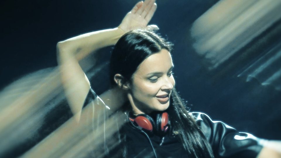 Miniatura aftermovie Korolova & Camelphat: DJ w czerwonych słuchawkach z uniesioną ręką w niebieskim świetle